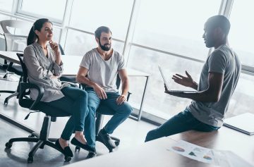 Group of attractive multiracial laughing businesspeople talking and joking sitting on chairs in light modern office, friendly multi-ethnic business team having fun, discussing business issues at meeting